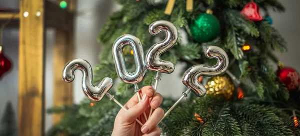 Bir Noel ağacının arka planındaki çubuklarda 2022 gümüş numaralarını tutan eller. Yeni yıl kavramı, tebrik kartları, şenlikli atmosfer..