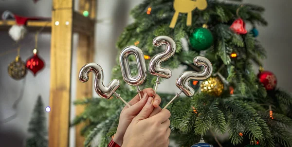 Bir Noel ağacının arka planındaki çubuklarda 2022 gümüş numaralarını tutan eller. Yeni yıl kavramı, tebrik kartları, şenlikli atmosfer..