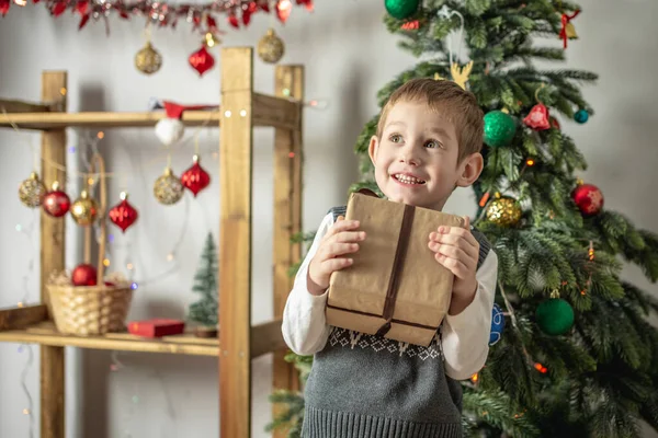 小さな男の子がお祝いの部屋でクリスマスツリーの隣に立っていて 彼の手にギフトボックスを持っています 休日の雰囲気と新年の気分の概念 — ストック写真