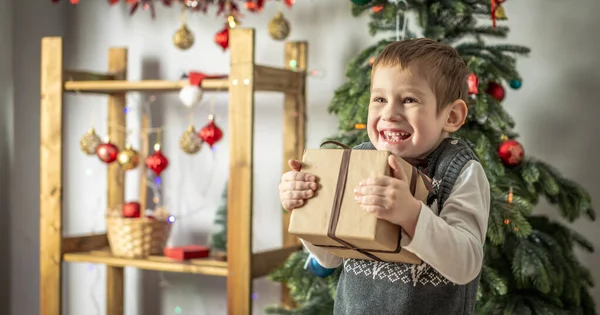 小さな男の子がお祝いの部屋でクリスマスツリーの隣に立っていて 彼の手にギフトボックスを持っています 休日の雰囲気と新年の気分の概念 — ストック写真