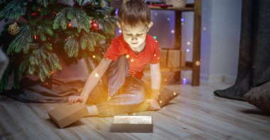 Küçük çocuk bir Noel ağacının yanında oturuyor ve sihirli bir ışık ve parlayan bir hediye kutusu açıyor. Yeni yıl havası ve şenlikli atmosfer kavramı.