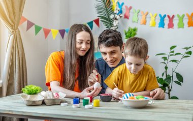 Anne, baba ve küçük oğlu parlak kıyafetler içinde dekore edilmiş bir odada yumurta boyuyorlar. Paskalya için aile hazırlığı kavramı, neşeli bahar havası.