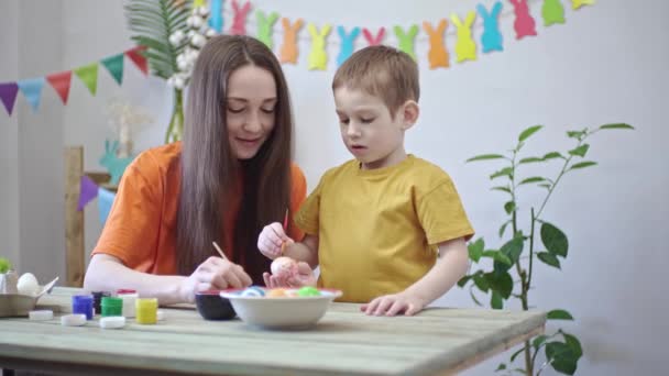 Η μαμά και ένα παιδί με φωτεινά ρούχα μαζί βάφουν αυγά σε ένα διακοσμημένο δωμάτιο. Έννοια της οικογενειακής προετοιμασίας για το Πάσχα, εορταστική ανοιξιάτικη διάθεση — Αρχείο Βίντεο