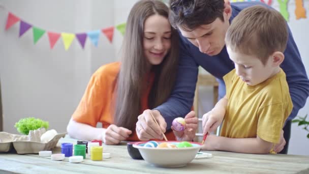 Mãe, pai e filho em roupas brilhantes estão colorindo ovos em uma sala decorada. Conceito de preparação familiar para a Páscoa, clima festivo de primavera — Vídeo de Stock