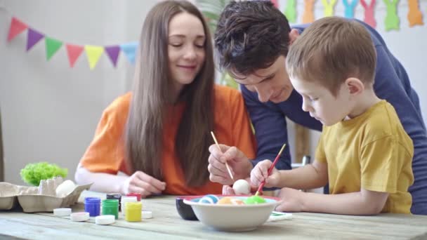 밝은 옷을 입은 엄마 아빠와 어린 아들은 장식된 방에서 달걀에 색칠을 하고 있다. 가족 이 부활절 준비를 하는 개념, 축제의 봄 분위기 — 비디오