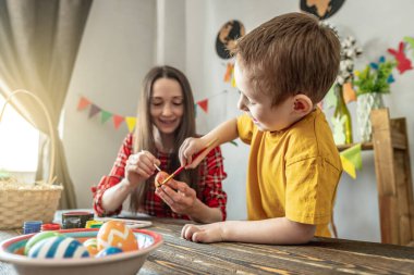 Anne ve çocuk birlikte parlak giysiler içinde dekore edilmiş bir odada yumurta boyuyorlar. Paskalya için aile hazırlığı kavramı, neşeli bahar havası.