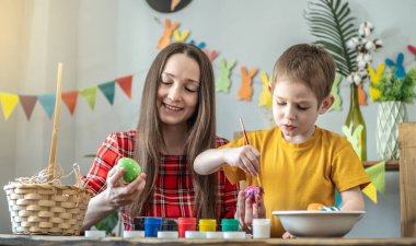 Anne ve çocuk birlikte parlak giysiler içinde dekore edilmiş bir odada yumurta boyuyorlar. Paskalya için aile hazırlığı kavramı, neşeli bahar havası.