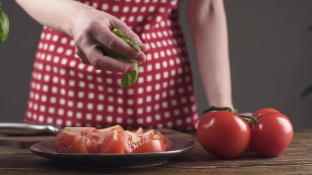 Na drewnianym stole znajduje się garnek z bazylią i talerz z pokrojonymi pomidorami. Kucharka w czerwonym fartuchu posypuje pomidory liśćmi bazylii. Szybko i zdrowo wiosenna sałatka witaminowa — Wideo stockowe