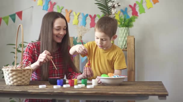 Mamãe e uma criança estão pintando ovos juntos em um quarto decorado para o feriado e se divertindo. Conceito de preparação para a Páscoa, clima de primavera e família feliz — Vídeo de Stock