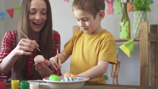 Mamãe e uma criança estão pintando ovos juntos em um quarto decorado para o feriado e se divertindo. Conceito de preparação para a Páscoa, clima de primavera e família feliz — Vídeo de Stock