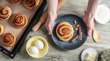 Masanın üzerinde taze tarçınlı çörekler, aşçıları için malzemeler ve tabakta iştah açıcı bir çörek var. Lezzetli ev yapımı hamur işleri ve rahat bir atmosfer anlayışı. Üst görünüm.