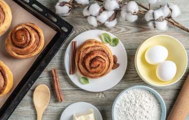 Masanın üzerinde taze tarçınlı çörekler, aşçıları için malzemeler ve tabakta iştah açıcı bir çörek var. Lezzetli ev yapımı hamur işleri ve rahat bir atmosfer anlayışı. Üst görünüm.