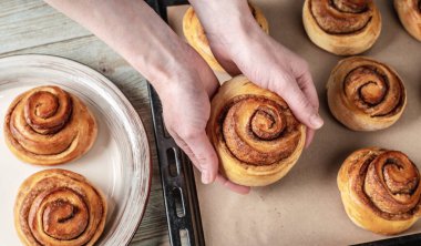 Dişi eller taze tarçınlı ruloları pişirme tepsisinden tabağa dikkatlice taşıyor. Lezzetli ev yapımı hamur işleri ve sıcak bir atmosfer kavramı. Üst görünüm.
