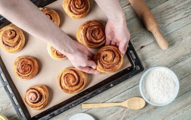 Kadın elleri dikkatle fırın tepsisinden taze tarçınlı çörekleri çıkarıyor. Lezzetli ev yapımı hamur işi ve rahat atmosfer kavramı.