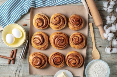 Tarçınlı çörekler, hazırlıkları için gerekli malzemeler ve mutfak gereçleri kağıt üzerindeki ahşap bir masaya konur. Lezzetli ev yapımı hamur işleri ve rahat bir atmosfer anlayışı. Üst görünüm.