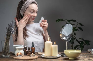 Kadın aynaya bakıyor ve guaj masajcısıyla masaj yapıyor. Mumlar ve masa üzerinde gerekli yağları olan aroma lambası. Yüz bakımı, aromaterapi, sabah akşam ritüelleri.