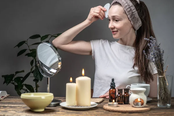 女は鏡を見てガッシュマッサージでマッサージをしている テーブルの上にエッセンシャルオイルとキャンドルとアロマランプ 顔のケア アロマセラピー 朝と夜の儀式の概念 ストック画像