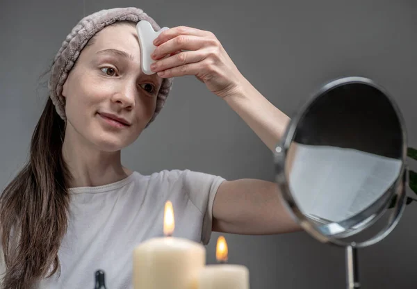 女は鏡を見てオイルでガッシュスクレーパーマッサージをしている テーブルの上にはロウソクが灯っている 顔のケア アロマセラピー 朝と夜の儀式の概念 ストック写真