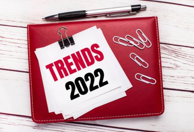 Hafif ahşap bir arka planda, bordo bir kalem ve defter. Not defterinde beyaz ataçlar ve üzerinde TRENDS 2022 yazılı beyaz kağıt var.