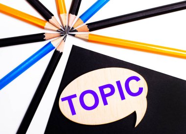 Çok renkli kalemlerin yanında siyah bir arkaplan üzerinde TOPIC yazılı tahta kart.