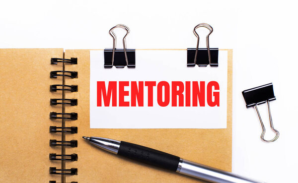 On a light background, a brown notebook with a pen, black clips and a white card with the text MENTORING