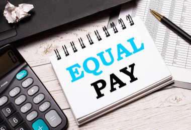 Açık renkli bir ofis masasında bir bilgisayar, siyah bir hesap makinesi, bir kalem ve EQUAL PAY yazılı bir defter vardır. İş konsepti.
