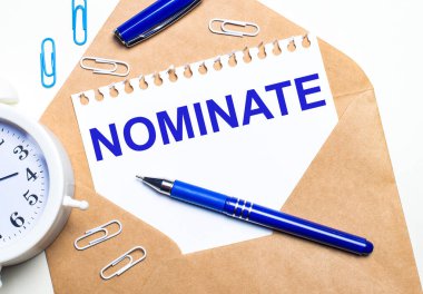 Hafif bir arkaplanda, bir el işi zarfı, bir çalar saat, ataçlar, mavi bir kalem ve üzerinde NOMINATE yazan bir kağıt.