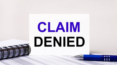 Açık gri arka planda, bir defter, mavi bir kalem ve CLAIM DENIED yazılı bir kağıt. İş konsepti