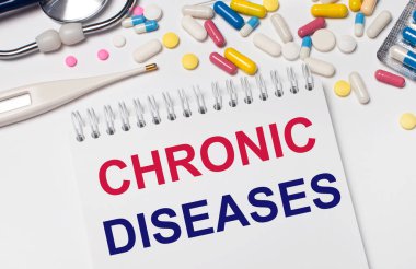 Açık bir arka planda, çok renkli haplar, steteskop, elektronik termometre ve CHRONIC DISEASES yazılı bir defter. Tıbbi konsept.