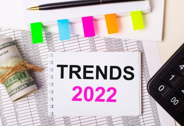 Hafif bir arka planda, bir kalem, dolarlar, TRENDS 2022 yazılı bir defter ve parlak çıkartmalar. İş konsepti
