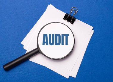 Mavi arka planda, siyah bir ataçın altında beyaz çarşaflar ve üzerinde AUDIT yazılı bir büyüteç.