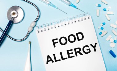 Açık mavi arka planda, bir elektronik termometre, bir steteskop, beyaz ve mavi haplar, şırıngalar ve FOOD ALLERGY yazılı bir defter. Tıbbi konsept