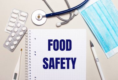 Açık gri arka planda, açık mavi tek kullanımlık yüz maskesi, bir stetoskop, bir elektronik termometre, haplar, bir kalem ve üzerinde Gıda Güvenliği yazan bir defter. Tıbbi konsept