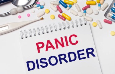 Açık bir arka planda, çok renkli haplar, bir steteskop, bir elektronik termometre ve PANIC DISORDER yazılı bir defter. Tıbbi konsept.