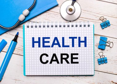 Hafif ahşap bir arka planda, steteskop, mavi bir not defteri, mavi ataç, mavi bir keçeli kalem ve HEALTH CARE yazılı bir kağıt. Tıbbi konsept