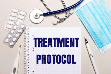 Açık gri arka planda, açık mavi tek kullanımlık yüz maskesi, bir stetoskop, bir elektronik termometre, haplar, bir kalem ve üzerinde TREATMENT PROTOCOL yazan bir defter. Tıbbi konsept