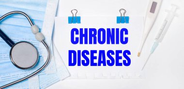 Hafif bir arka planda bir stetoskop, bir elektronik termometre, bir şırınga, bir yüz maskesi ve CHRONIC DISEASES yazılı bir kağıt parçası bulunur. Tıbbi konsept