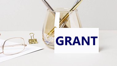 İş yerinde ve gri masada altın gözlük, ataç, bardaktaki kalemler ve içinde GRANT yazan beyaz kart var. Merkez ofis. İş yeri yakın çekim