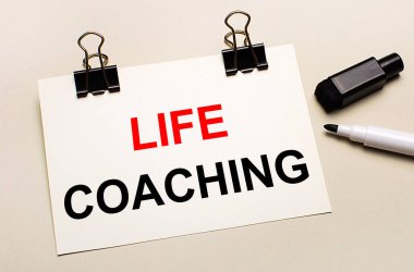 Açık bir arkaplanda, siyah bir keçeli kalem ve siyah ataçlar üzerinde LIFE COACHING metni olan beyaz bir kağıt parçası