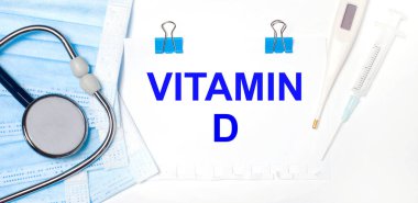 Hafif bir arka planda bir stetoskop, bir elektronik termometre, bir şırınga, bir yüz maskesi ve VITAMIN D yazılı bir kağıt parçası bulunur.