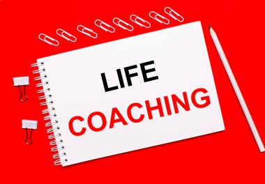 Parlak kırmızı bir arka planda, beyaz bir kalem, beyaz ataçlar ve üzerinde LIFE COACHING yazan beyaz bir defter.