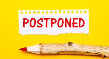 Parlak sarı bir arka planda, büyük bir tahta kalem ve üzerinde POSTPONED yazan yırtık bir kağıt parçası.