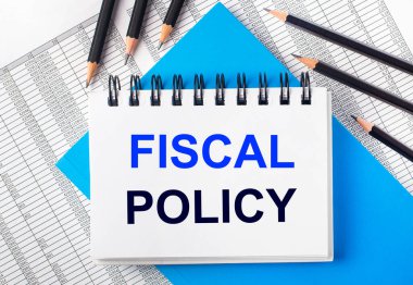 Masanın üzerinde FISCAL POLICY yazılı beyaz defter mavi arka planda siyah kalemlerin yanında ve raporlar. İş konsepti