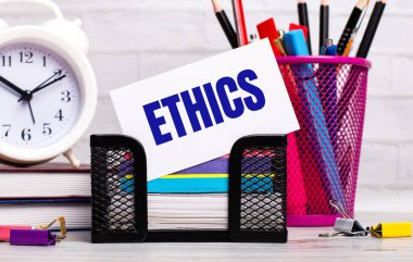 Ofis masasında günlükler, bir çalar saat, kırtasiye malzemeleri ve ETHICS yazılı beyaz bir kart var. İş konsepti.