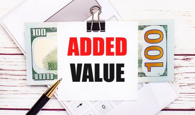 Hafif ahşap bir masanın üzerinde beyaz bir hesap makinesi, bir kalem, faturalar ve siyah bir ataçın altında ADDED VALUE yazılı bir kağıt var. İş konsepti