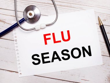 Hafif ahşap bir masanın üzerinde steteskop, bir kalem ve üzerinde FLU SEASON yazılı bir kağıt var. Tıbbi konsept