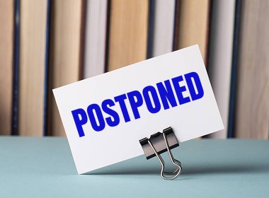 Üzerinde POSTPONED yazan beyaz bir kart, kitapların arka planına karşı masada duran kağıtlar için bir şarjör üzerinde duruyor. Odaklanmayın