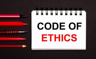 Siyah arkaplanda kırmızı kalemler, kalemler ve işaretleyicilerin yanında ETHICS 'in metin kodunu içeren beyaz bir defter.