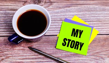 Ahşap bir masanın üzerinde - içinde kahve, kalem ve renkli parlak çıkartmalar olan mavi bir fincan My Story. Yukarıdan görüntüle