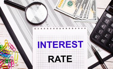 Ofis masasında içinde INTEREST RATE yazan bir defter, bir kalem, bir hesap makinesi, dolar, renkli ataçlar ve bir büyüteç var. Şık bir iş yeri. İş konsepti